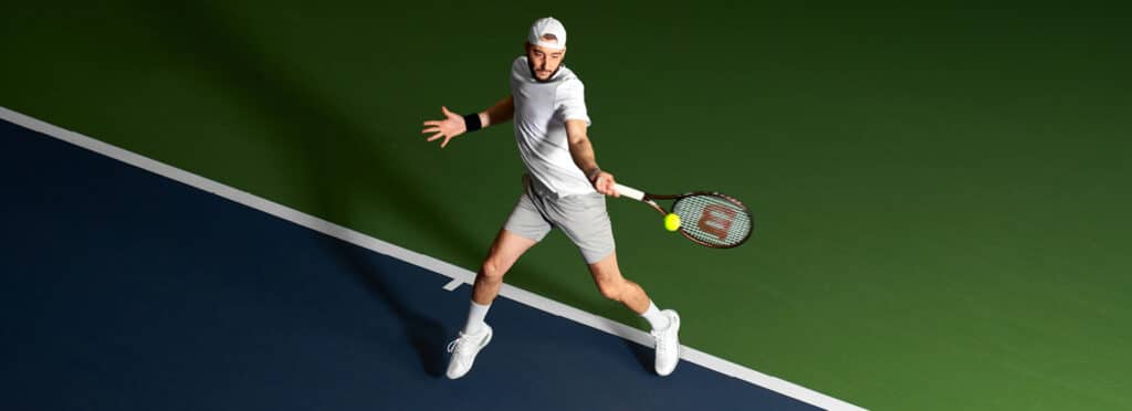A tennis player dressed in all white, including a cap, shirt, shorts, and sneakers, is actively swinging a racket with both hands to strike a yellow tennis ball. The action takes place on a court divided into green and blue sections, with the player positioned near the dividing line.