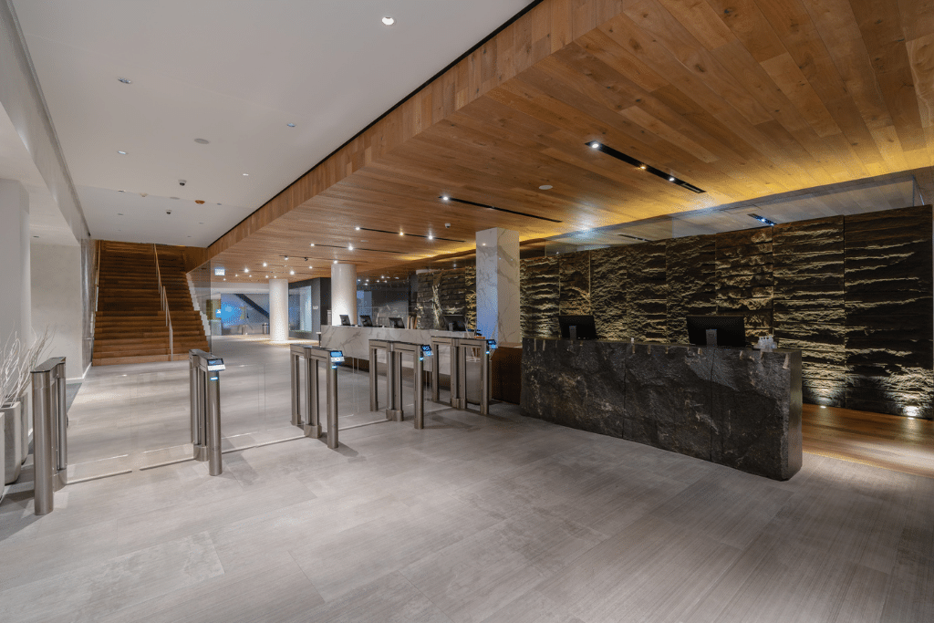 Modern and spacious lobby area featuring a wooden ceiling, stone walls, and large gray tiled flooring. The space includes access control turnstiles, a stone reception desk, and a staircase leading to an upper level. Warm lighting enhances the welcoming atmosphere.