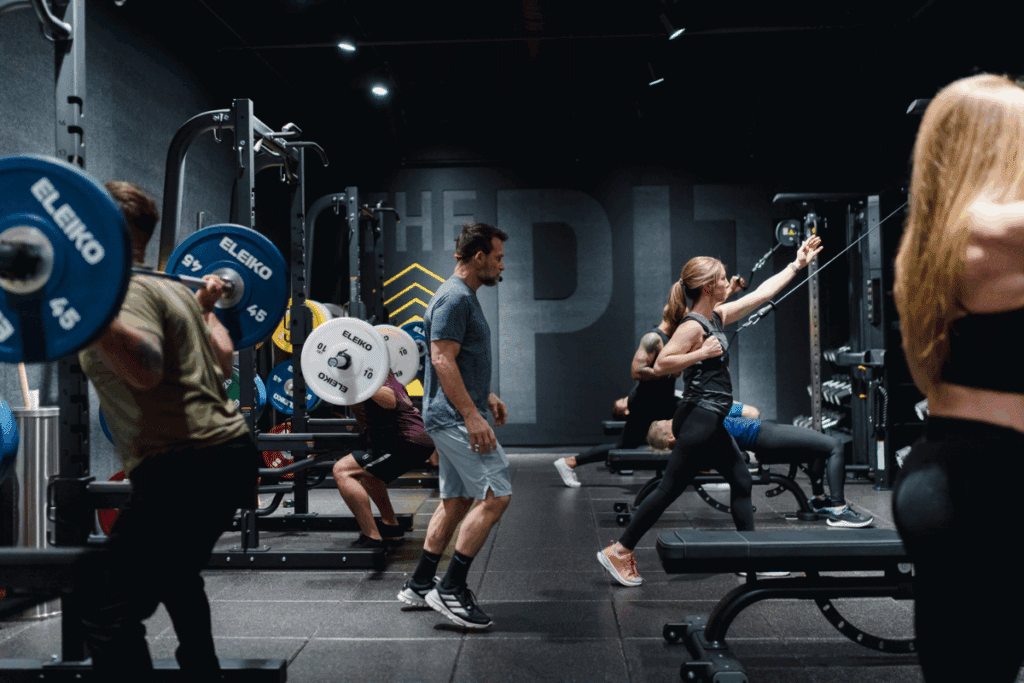 Gym environment with several individuals performing different exercises among weightlifting racks, benches, and various gym equipment. The space is labeled 'THE PIT' with bold text on the back wall, emphasizing a high-intensity training area.