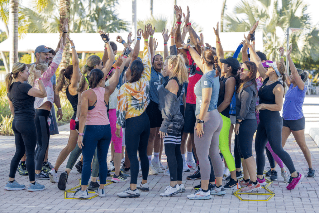 Outdoor fitness or team-building activity at Midtown Athletic Club with participants raising hands in a circle, set against a backdrop of palm trees.
