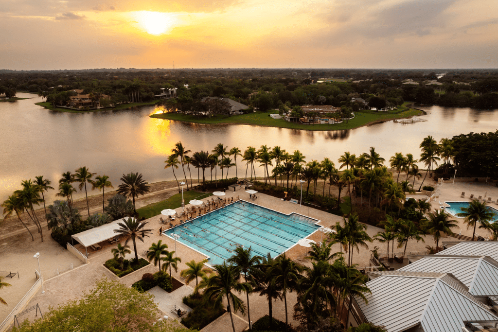 Scenic resort-style pool area at Midtown Athletic Club featuring lounge chairs, umbrellas, palm trees, and a tranquil lake view at sunset.