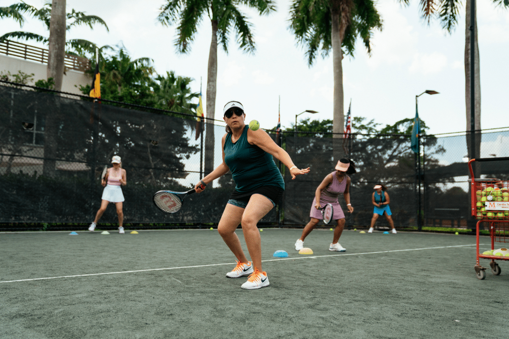 Group of four people playing tennis on an outdoor court. One player in the foreground is preparing to hit a tennis ball with a racket. The court is enclosed by a black chain-link fence, with palm trees in the background and a cart filled with tennis balls visible on the right side.