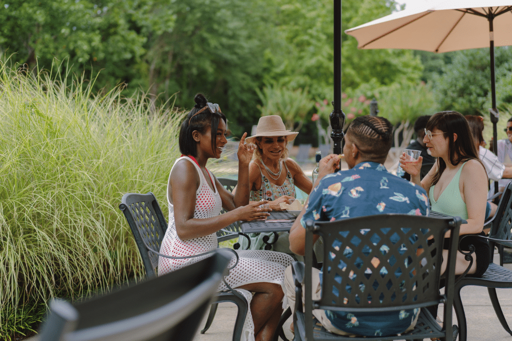 Group of people seated around an outdoor table, engaged in conversation and enjoying drinks. The setting features surrounding greenery and a large umbrella providing shade, creating a relaxed and social atmosphere.