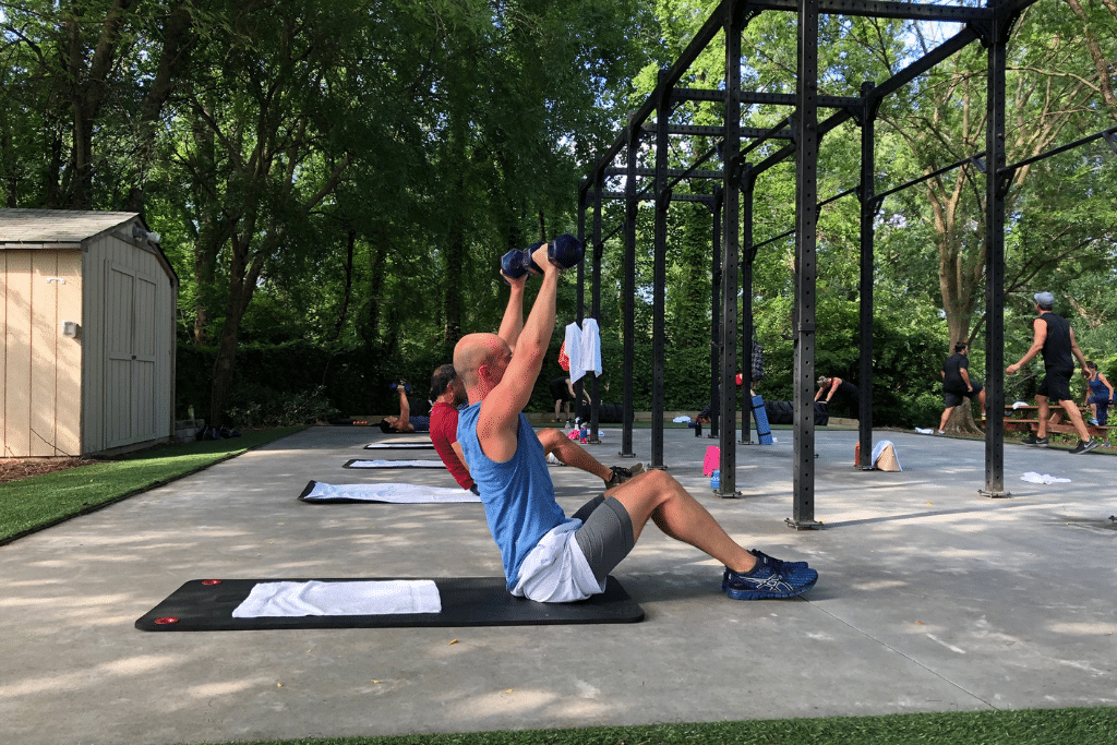 Outdoor exercise area surrounded by trees, featuring several people engaged in various workouts. In the foreground, a person wearing a blue sleeveless shirt and gray shorts sits on a black exercise mat, lifting two dumbbells overhead. In the background, others are using a metal bar structure for exercises like pull-ups, creating a dynamic, nature-integrated fitness environment