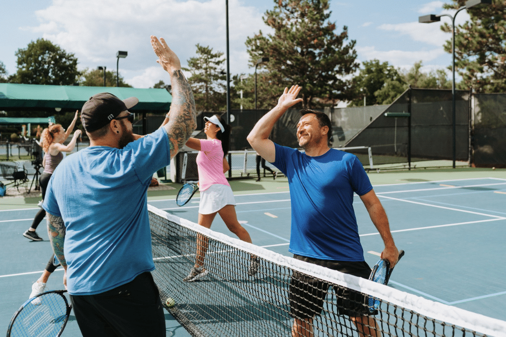 Four people on an outdoor tennis court, with two players in the foreground giving each other a high-five over the net while holding tennis rackets. Both are dressed in blue shirts, and one wears a cap. In the background, another player in a pink shirt and white cap stands near the net, while a fourth person is further back near green structures. The court is surrounded by trees under a clear sky.