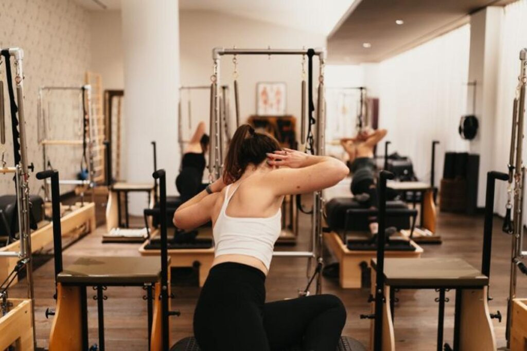 Woman performing Pilates on a reformer machine at a boutique Pilates studio near Palatine, near Buffalo Grove and Schaumburg. The bright fitness studio features multiple reformer machines, wooden floors, and large windows—ideal for athletic reformer Pilates and mat Pilates classes.