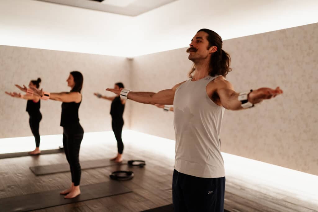 Group Pilates class in a bright modern studio with participants performing standing poses with arms extended on yoga mats.