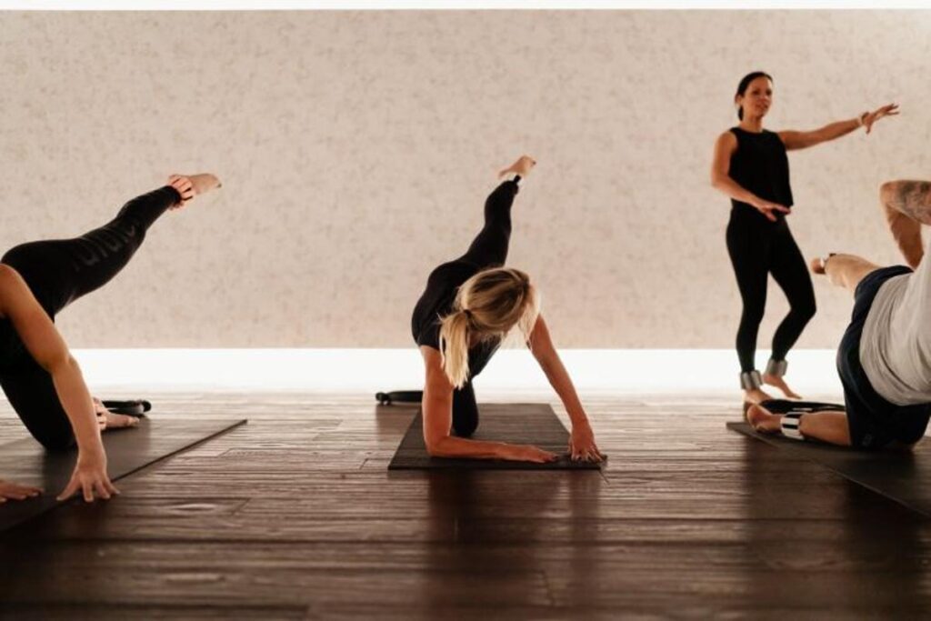 A group of people in a fitness studio performing a balance pose on yoga mats, each extending one arm and the opposite leg. An instructor stands in the background, observing and guiding the class. The room features wooden flooring and a light-colored wall with subtle patterns.