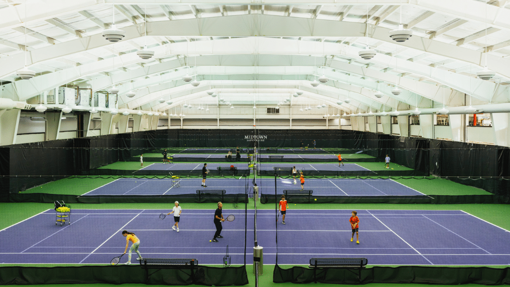 Indoor tennis courts at Midtown Athletic Club Montreal featuring six blue and green courts under a high arched ceiling, with members actively playing and receiving coaching in a spacious, well-lit environment.
