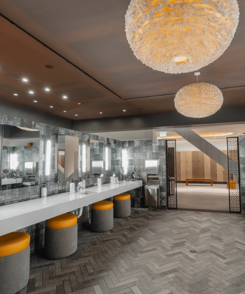 chi locker arc Modern locker room featuring large, round textured light fixtures and recessed ceiling lighting. A long white countertop with multiple sinks and mirrors lines one wall, each sink accompanied by a soap dispenser. Yellow-cushioned cylindrical stools are placed beneath the counter. The floor is covered in herringbone-patterned tiles, and an open doorway in the background reveals a room with wooden lockers and a bench.