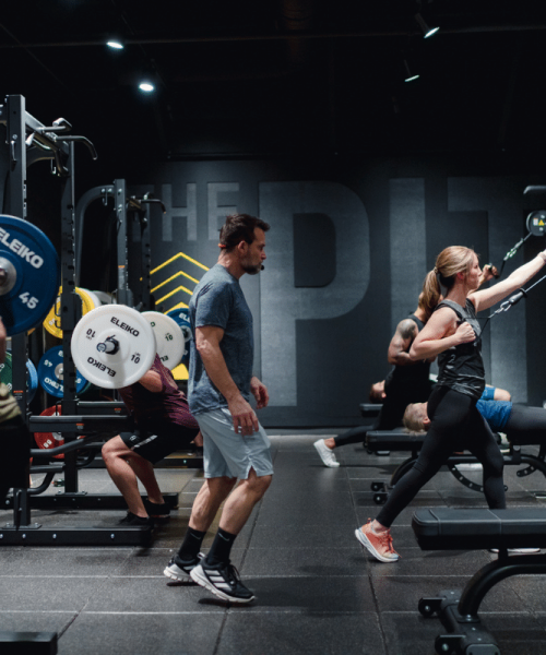 Gym environment with several individuals performing different exercises among weightlifting racks, benches, and various gym equipment. The space is labeled 'THE PIT' with bold text on the back wall, emphasizing a high-intensity training area.