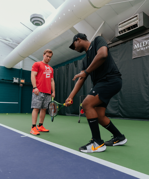 chi tennis (500×600) Two individuals on an indoor tennis court, with one person in a black outfit and cap demonstrating a tennis stroke using a racket and ball. The other person, wearing a red shirt and gray shorts, stands nearby observing. The court features netting in the background.