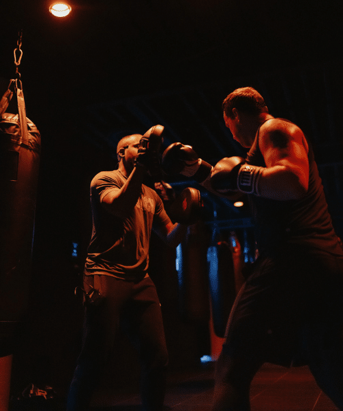 generic groupfitness Two individuals in a dimly lit boxing gym engaged in a training session. One person holds up focus mitts while the other, wearing boxing gloves, practices punches. Warm overhead lighting casts a dramatic glow on the scene, and several heavy punching bags hang in the background, contributing to the gritty, focused atmosphere.