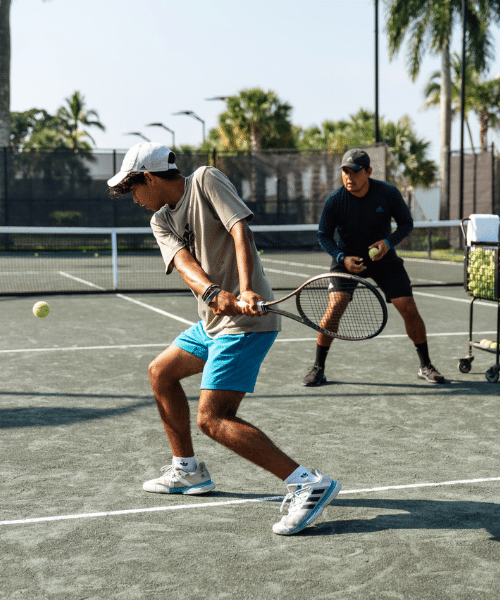 WES_Tennis_Outdoor_GreenClay_PrivateCoaching_Backhand_500x600 Outdoor tennis lesson at Midtown Athletic Club with a player preparing to hit a ball while receiving coaching support, set against a backdrop of palm trees and fenced court.