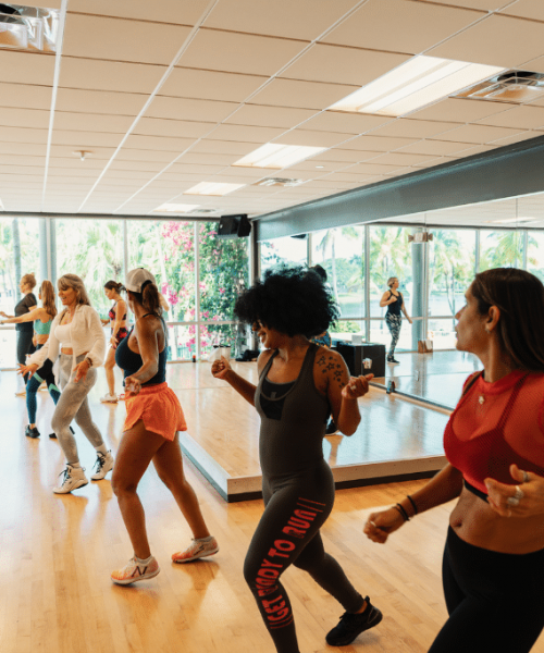 WES_GroupZ_Zumba_1024x683 Energetic group fitness class at Midtown Athletic Club with members participating in dance-based aerobic exercises in a mirrored, sunlit studio with wood flooring.