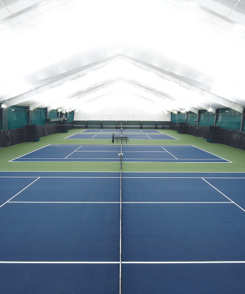 A well lit room houses 4 blue tennis courts.