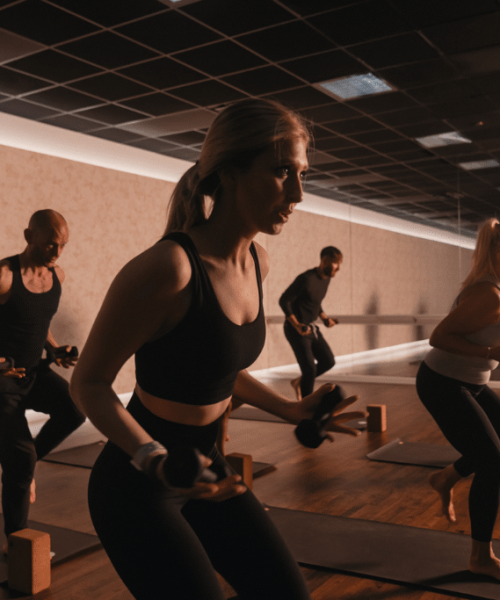 5 men and women hold small weights and lift one leg off the floor as they look ahead.
