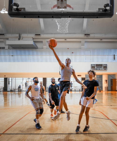 Group of people playing basketball on an indoor court. One player is mid-air near the hoop, attempting a layup or dunk, while others are positioned around the court, either defending or observing. The background features a scoreboard and gym equipment.