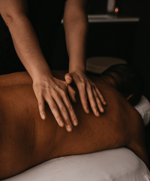 Person lying face down on a massage table receiving a back massage. The masseuse's hands are applying pressure to the upper back, and the dim lighting creates a calm, relaxing atmosphere.