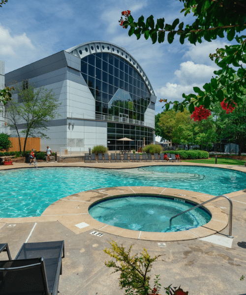 Outdoor swimming pool area with a hot tub in the foreground, surrounded by a concrete deck with lounge chairs and greenery. A modern building with large glass windows and an arched roof stands in the background. People, including children, are enjoying the sunny day around the pool under a partly cloudy sky.