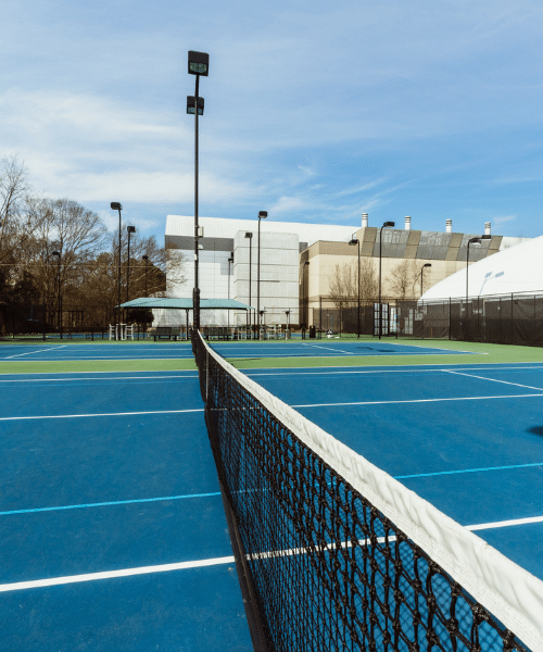 Here’s a descriptive alt text for the twenty-third image: "Outdoor tennis court with a blue playing surface and white boundary lines. A net stretches across the center of the court, and tall light poles surround the area. In the background, buildings and trees are visible under a partly cloudy sky.